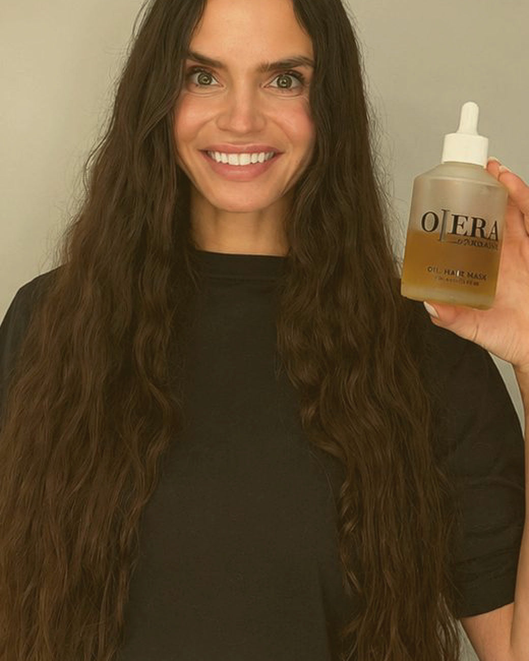 Founder after: long, smooth, healthy wavy hair, holding a bottle of OLERA